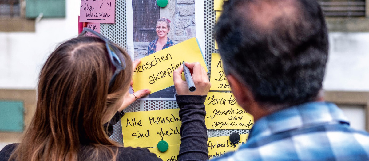 Eine junge Frau und ein älterer Mann vor einer Stele mit vielen Zetteln. Die Frau schreibt gerade einen neuen Zettel mit der Aufschrift "Menschen akzeptieren"