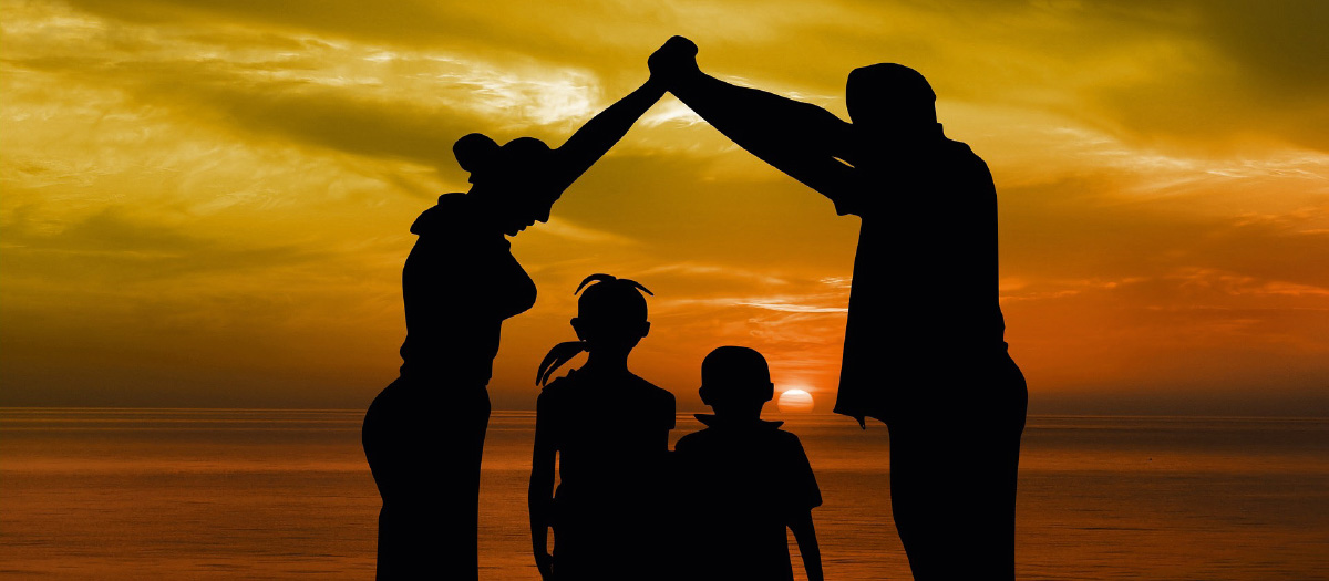 Zwei Erwachsene und zwei Kinder, die vor einem Sonnenuntergang stehen. Die Erwachsene halten schützend ihre Hände über die beiden Kinder im Zentrum.