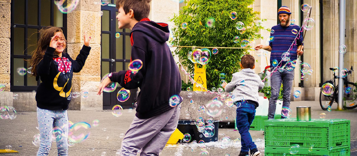 Ein Erwachsener macht vor einem Gebäude sehr viele Seitenblasen. Drei Kinder haben sichtlich Freude an den vielen Seifenblasen.