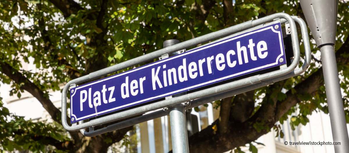 Straßenschild mit der Aufschrift "Platz der Kinderrechte" vor einem Baum mit grünen Blättern.